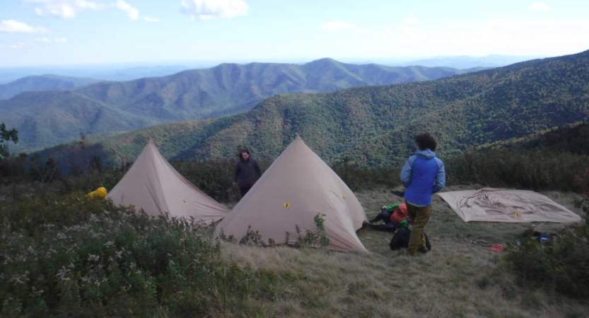 A person stands beside two tents set up in a green open area overlooking the vast Blue Ridge Mountains. 